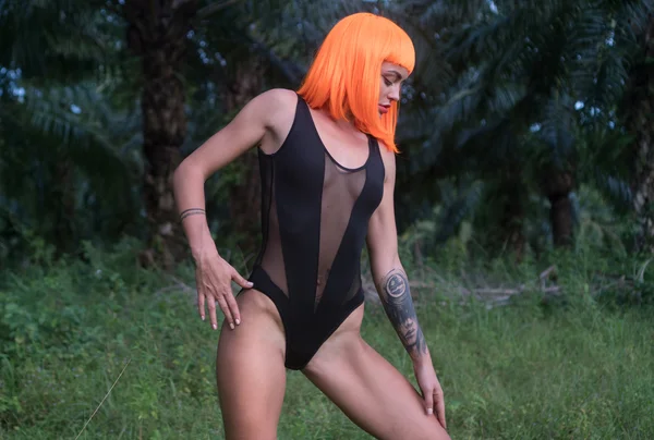 Sexy beautiful woman in modern futuristic style posing in palm trees forest. Creative look of tattooed woman wearing black bodysuit and orange wig
