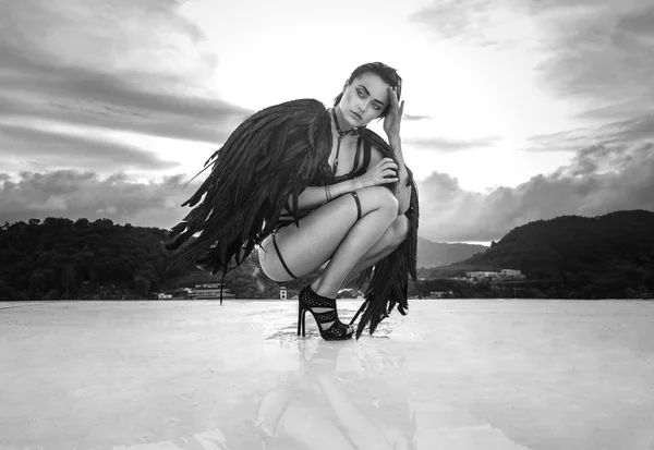 Black and white photo of beautiful seductive angel woman wearing lingerie and leather belts crouching on the roof over cloudy sky