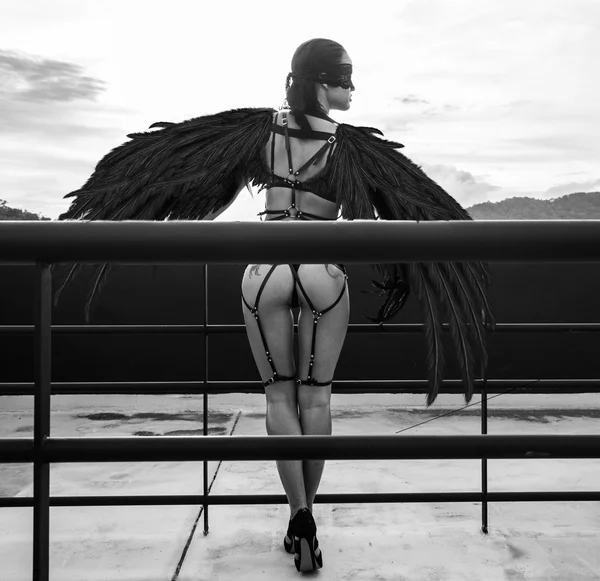 Back view black and white photo of beautiful seductive angel woman with covered eyes wearing lingerie and leather belts standing on the roof over cloudy sky