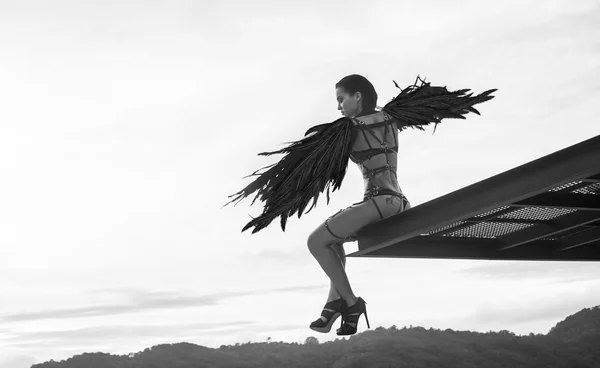 Black and white photo of beautiful seductive angel woman wearing lingerie and leather belts sitting on the roof edge with wind in her wings over cloudy sky