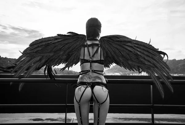 Back view black and white photo of beautiful seductive angel woman with covered eyes wearing lingerie and leather belts standing on the roof over cloudy sky