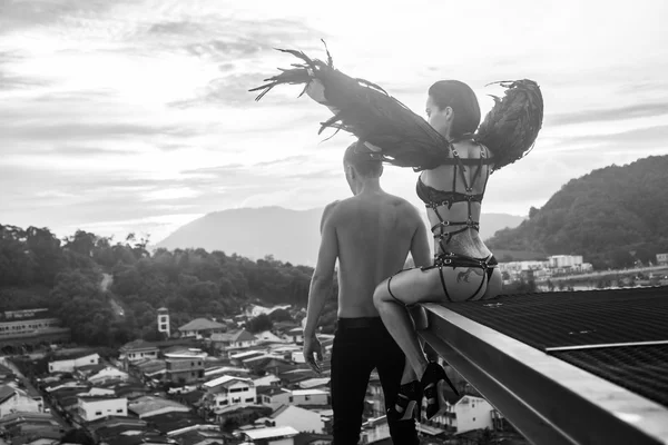 Black and white photo of romantic scene with shirtless man and sensual angel woman wearing lingerie, leather belts and high heels on the rooftop over sky and city landscape background