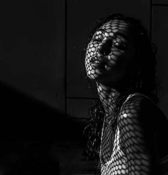 Black and white closeup portrait photo of beautiful seductive brunette woman posing beside the outdoor swimming pool during sunny summer day with shadow net reflection on her face and body