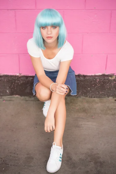 Sexy young girl in modern futuristic style with blue wig and cigarette crouching and looking into the camera over pink wall background