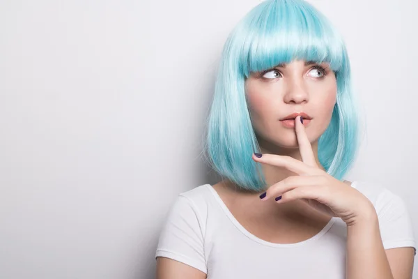 Closeup portrait of cheeky young girl in modern futuristic style wearing blue wig with curious expression and finger on lip while looking sideways over white wall background with copyspace