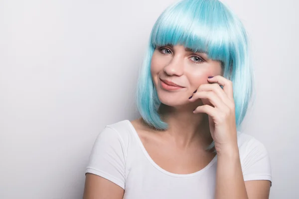 Cheeky young girl in modern futuristic style with blue wig smiling and looking into the camera over white wall background with copyspace