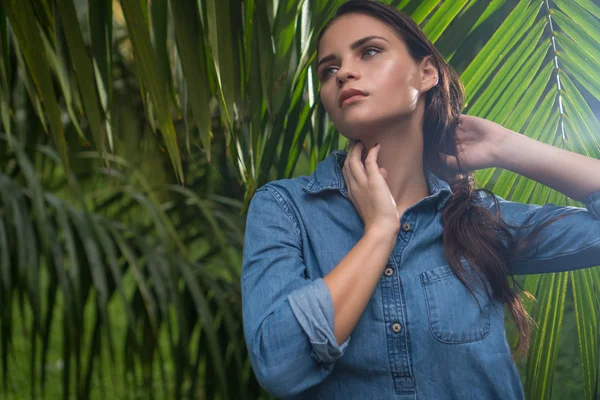 Closeup portrait of young brunette woman wearing jeans shirt in jungle during sunny day over palm trees background