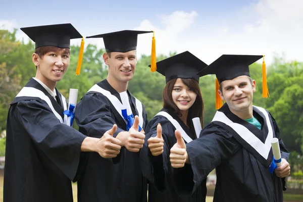 Happy students in graduation gowns showing diplomas with thumbs