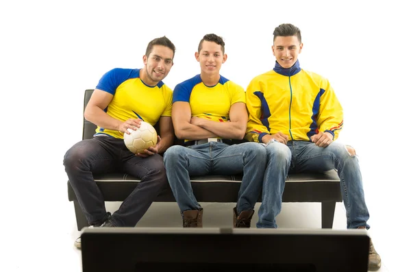 Three friends sitting on sofa wearing yellow sports shirts watching television with enthusiasm, white background, shot from behind tv