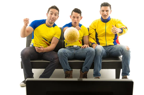 Three friends sitting on sofa wearing yellow sports shirts watching television with enthusiasm, golden ball flying in front, white background, shot from behind tv
