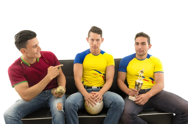 Three friends sitting on sofa wearing sports shirts smiling mocking interacting with each other holding trophy and ball, white background