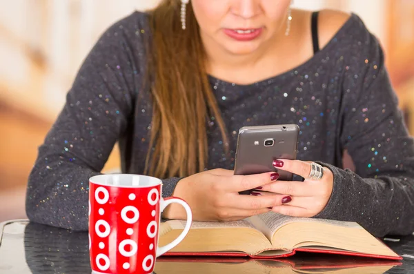 Young brunette woman wearing dark sweater sitting down pressing mobile phone with red mug in front, book lying on table