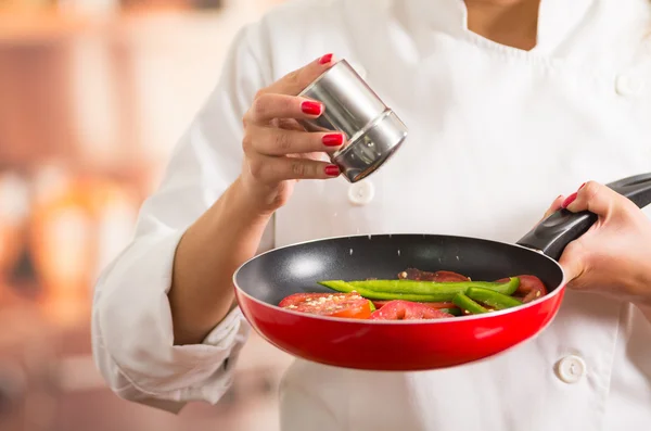 Closeup woman chefs hands holding red skillet with chopped vegetables inside, adding salt from metal container