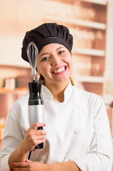 Woman chef wearing cooking outfit posing happily holding electric handheld blender