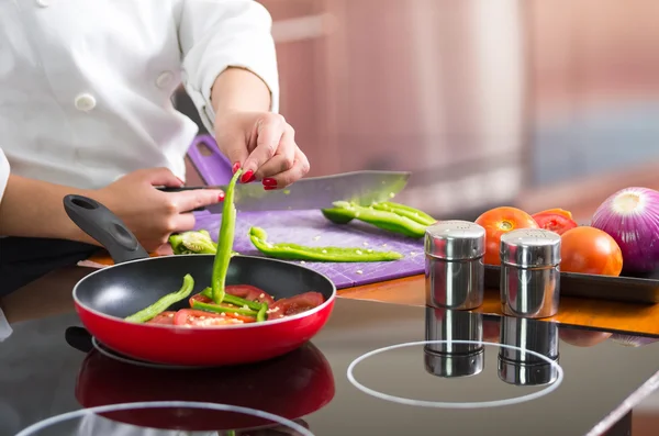Closeup chef cutting green peppers on wooden surface with red skillet in front frying up vegetables