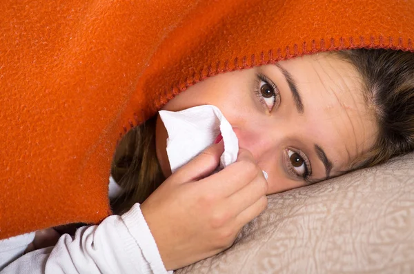 Brunette lying down under orange blanket and blowing her nose, sick with flu concept