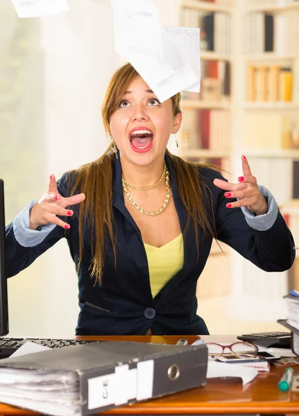 Office woman sitting by desk throwing papers up in the air and screaming
