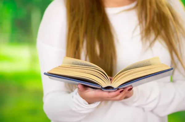 Closeup open book held by woman wearing white sweater and long hair visible, garden background