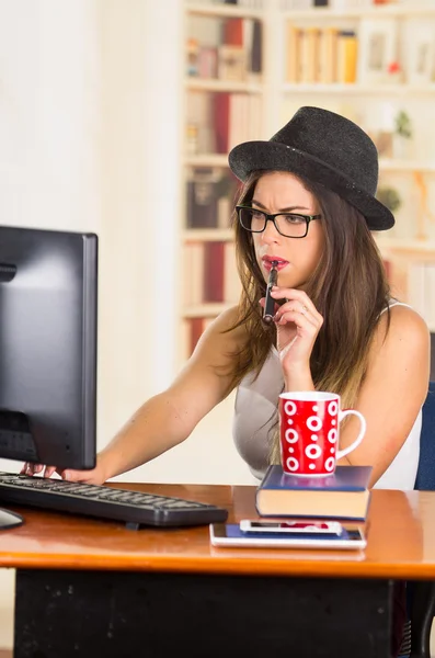 Young hip brunette office worker wearing glasses and fashionable hat, sitting down by desk with computer while smoking e-cigarette, red coffee mug placed on books