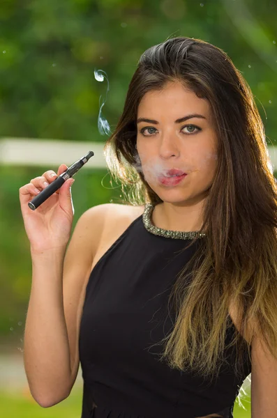 Classy brunette lady wearing black dress standing in garden environment smoking e-cigarette and posing for camera