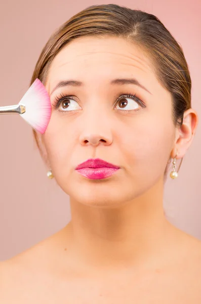 Headshot young pretty hispanic woman brunette with red lipstick holding makeup brush against side of face, serious facial expression, pink background