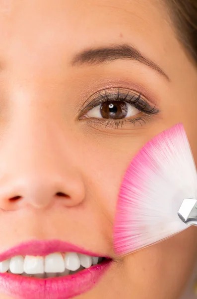 Closeup headshot young pretty hispanic woman holding pink white makeup brush up next to eye and smiling