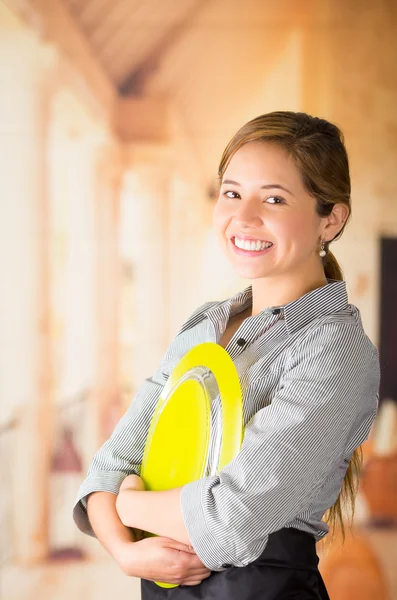Young brunette waitress holding large yellow plate to chest and smiling at camera, shot from profile angle