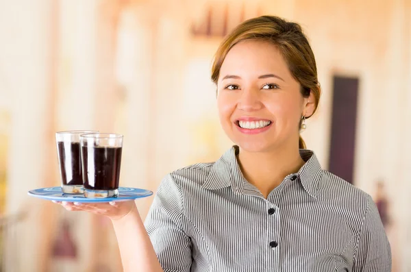 Young brunette waitress wearing uniform with friendly smile, holding up tray containing two glasses of dark liquid using one arm