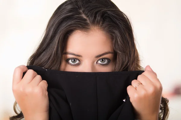 Headshot attractive brunette facing camera covering half her face with black clothing, white studio background