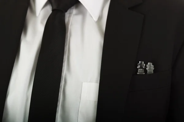 Closeup black suit jacket chest pocket with glass chess pieces sitting inside, white shirt and tie visible