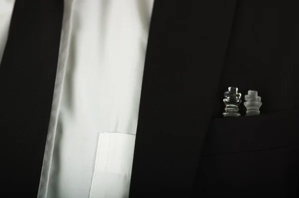 Closeup black suit jacket chest pocket with glass chess pieces sitting inside, white shirt and tie visible