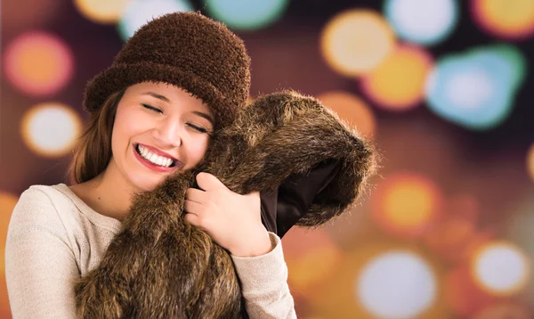 Pretty young brunette woman wearing fur style jacket, hat and scarf posing happily with a glamorous blurry light drops background