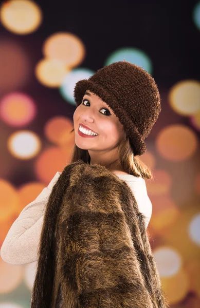Pretty young brunette woman wearing fur style jacket, hat and scarf posing happily with a glamorous blurry light drops background