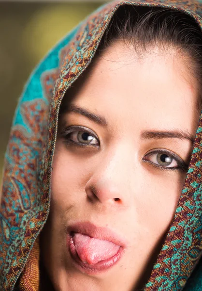 Closeup woman wearing blue, grey and brown coloured scarf covering head revealing face, holding tongue out