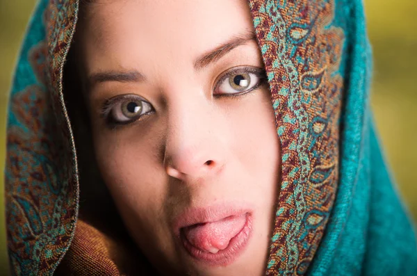 Closeup woman wearing blue, grey and brown coloured scarf covering head revealing face, holding tongue out