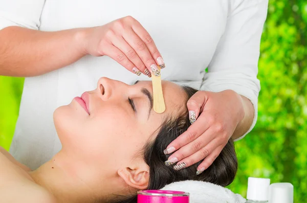 Closeup womans face receiving facial hair waxing treatment, hand using wooden stick to apply wax, beauty and fashion concept