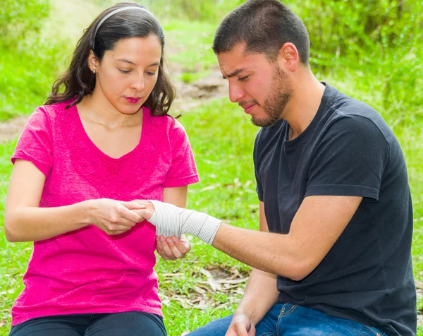 Young man with injured wrist sitting and getting bandage compression wrap from female, outdoors environment