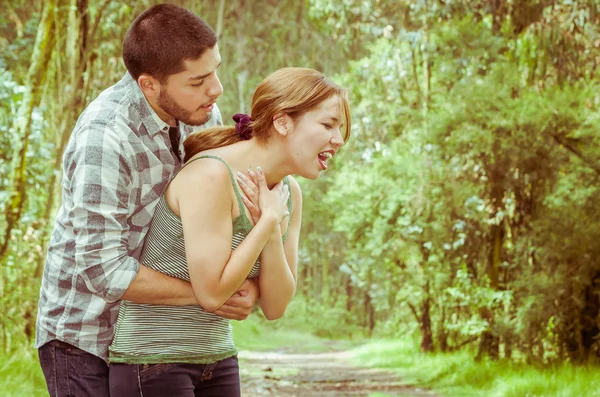 Young woman choking with man standing behind performing heimlich maneuver, park environment and casual clothes