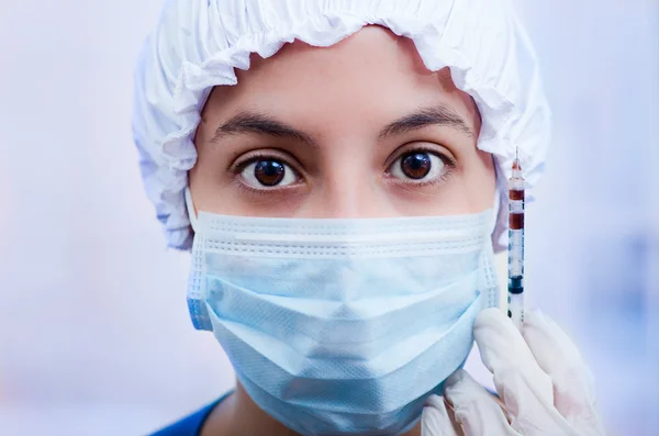 Closeup headshot nurse wearing bouffant cap and facial mask holding up syringe needle for camera