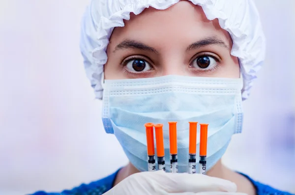 Closeup headshot nurse wearing bouffant cap and facial mask holding up syringes for camera