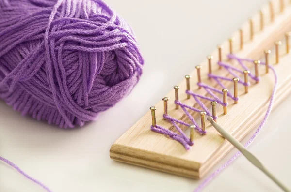Purple yarn ball  on white surface, loose end tied up to wooden board with nails, handcraft textile concept
