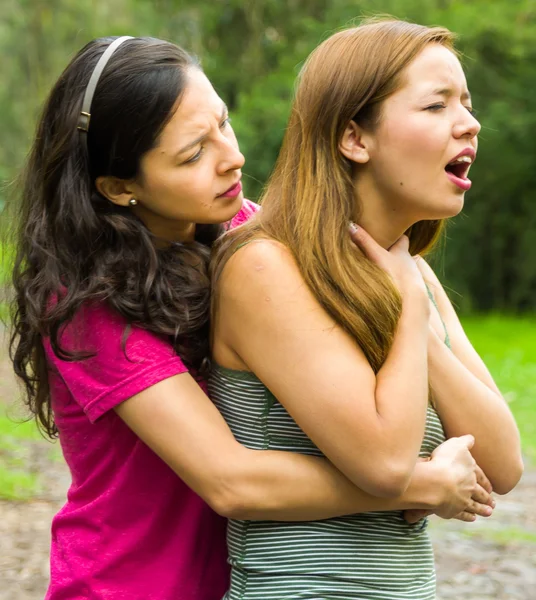 Young woman choking with lady standing behind performing heimlich maneuver, park environment and casual clothes