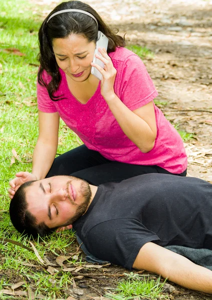 Young man lying down with medical emergency, woman sitting by his side calling for help, outdoors environment