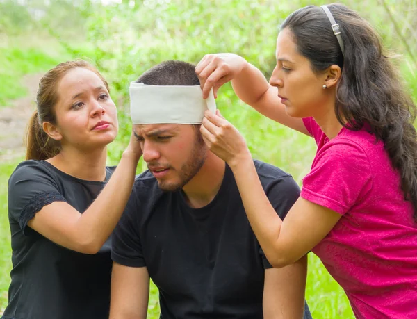Young man with head injury receiving treatment and bandage around skull from two women, outdoors environment