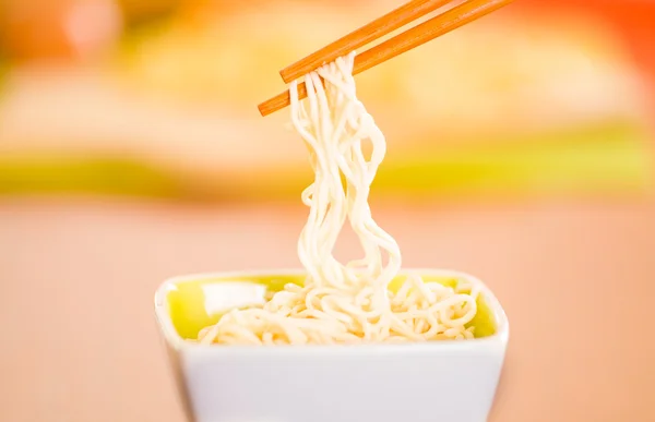 Elegant presentation of cooked noodles inside small square bowl sitting on wooden surface, sticks grabbing food, blurry background