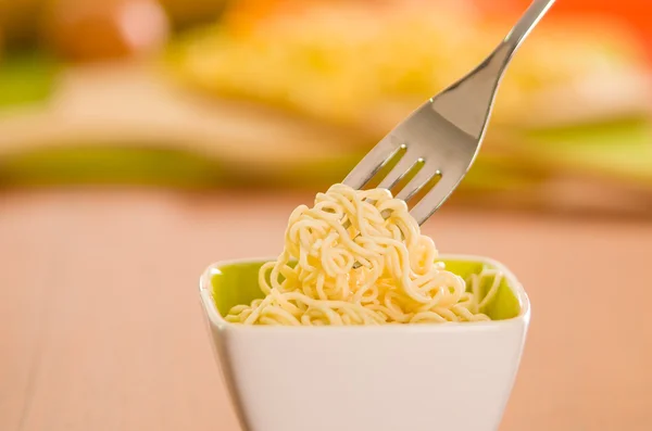 Elegant presentation of cooked noodles inside small square bowl sitting on wooden surface, fork grabbing food, blurry background