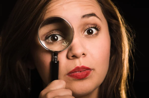 Young woman facing camera holding magnifying glass over right eye creating enhanced effect, black background