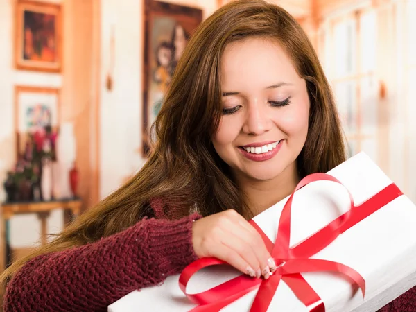 Brunette with beautiful smile holding up present happily posing for camera, white wrapping and red ribbon, household background