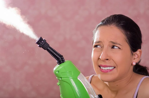 Brunette woman holding steam cleaner machine and vapor coming out, smiling to camera, pink background