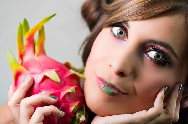 Headshot brunette, dark mystique look and green lipstick, holding up pink pitaya fruit, looking into camera